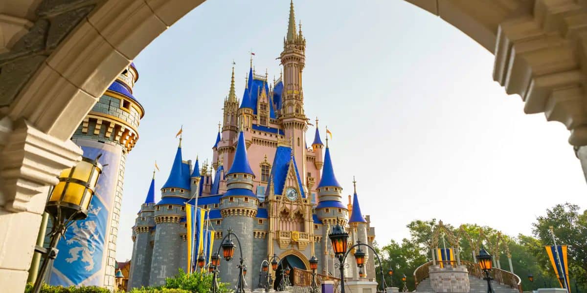 A photo of a large fairytale castle with blue and gold rooftops, seen through a stone archway on a sunny day. Decorative flags and vintage-style lamps line the walkway leading to the castle as Disney World crowds vanish from plane sight as Disney news is reported. Orlando tourism record