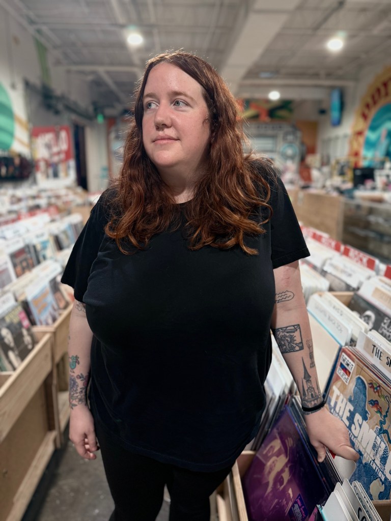 A medium portrait of an individual with long auburn hair and several tattoos on their arms, standing in the aisle of a record store. They are wearing a black t-shirt and looking off-camera, surrounded by bins of vinyl records with a "Rock" section sign visible in the background.