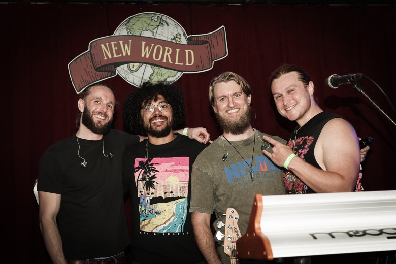 Four smiling individuals stand together for a group portrait in front of a dark red stage curtain. A large logo above them features a globe and a banner that reads "NEW WORLD." The person on the far right gives a hand gesture, while a white keyboard and the headstock of a bass guitar are visible in the foreground