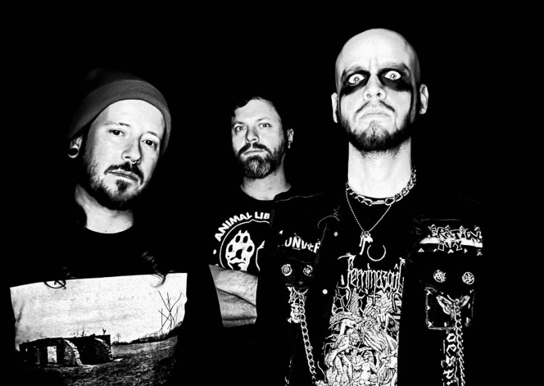 A trio of serious-looking musicians. The subject on the left wears a beanie and large ear gauges. The central person wears a dark t-shirt, and the person on the right wears a patched, chain-adorned vest and metal necklaces.