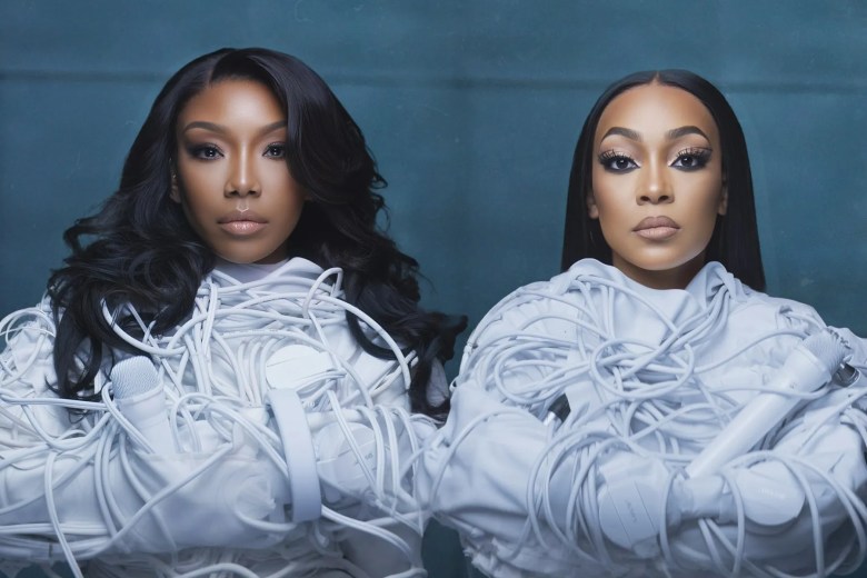 A dramatic portrait of two people with striking makeup, standing against a dark, slate-blue background. They are wearing clothing made of white coiling cords or cables, creating a sculptural, artistic effect.
