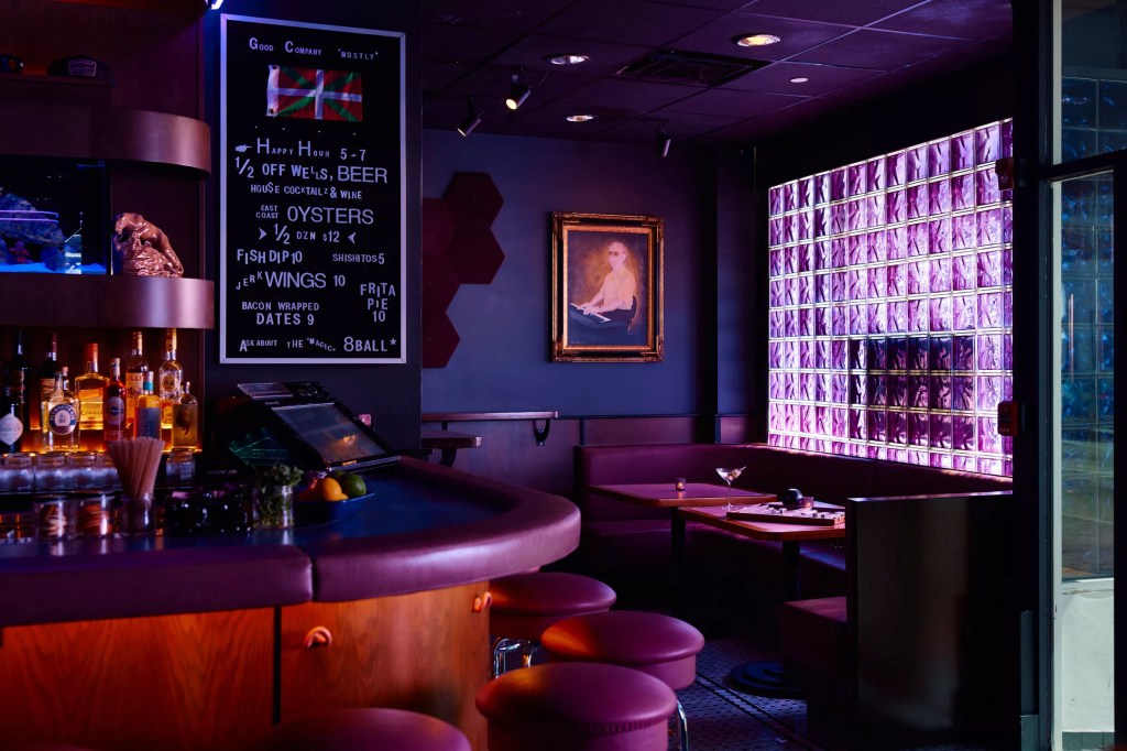 photo of the corner of a bar awash in purple lighting with a drink list on a chalkboard, empty seats at bar, and empty booths in the corner