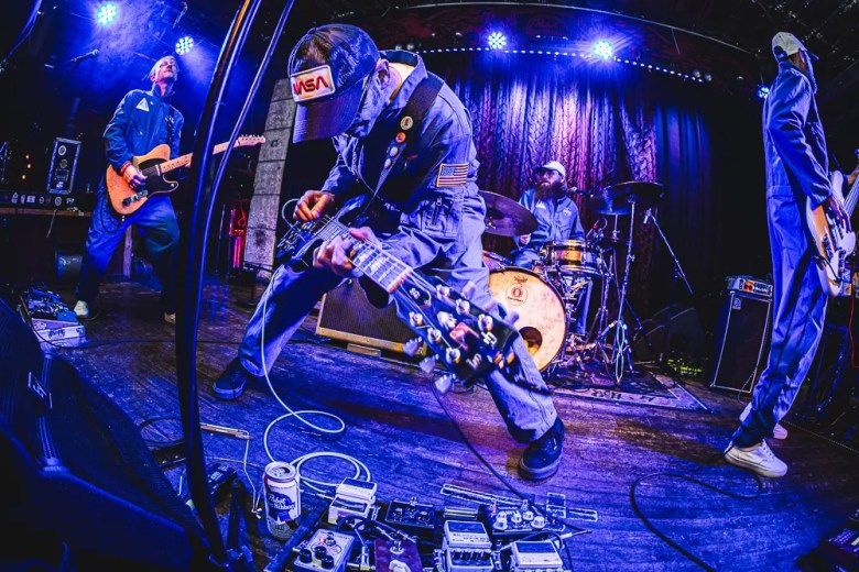 A musician in a jumpsuit and cap is captured playing an electric guitar aggressively in the foreground, bending over a stack of effects pedals. Blue stage lighting illuminates the drummer and another guitarist in the background.