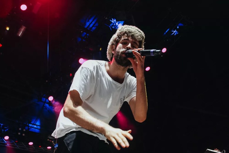 Jack Met of AJR performing live on stage, singing into a microphone, wearing a white t-shirt and his signature fuzzy hat under red and blue concert lighting.