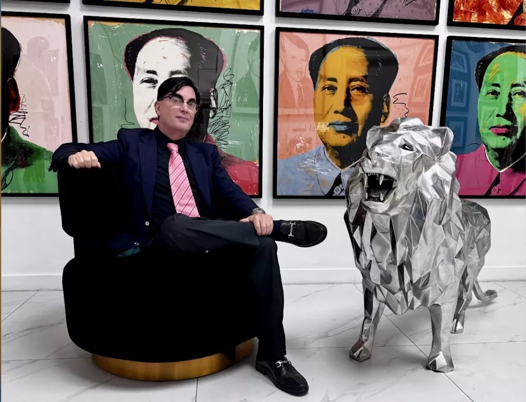 photo of a seated man with a conspicuous toupee beside a large lion figurine with ostensible Andy Warhol works in the background
