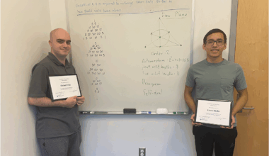 Photo: Two students stand on each side of a whiteboard with a math problem.