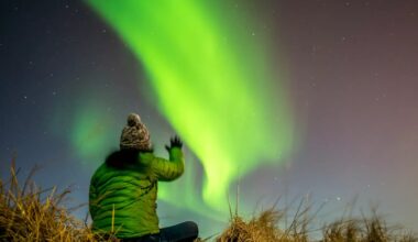 photo of a person in a beanie and thick green jacket looking up at green Northern Lights