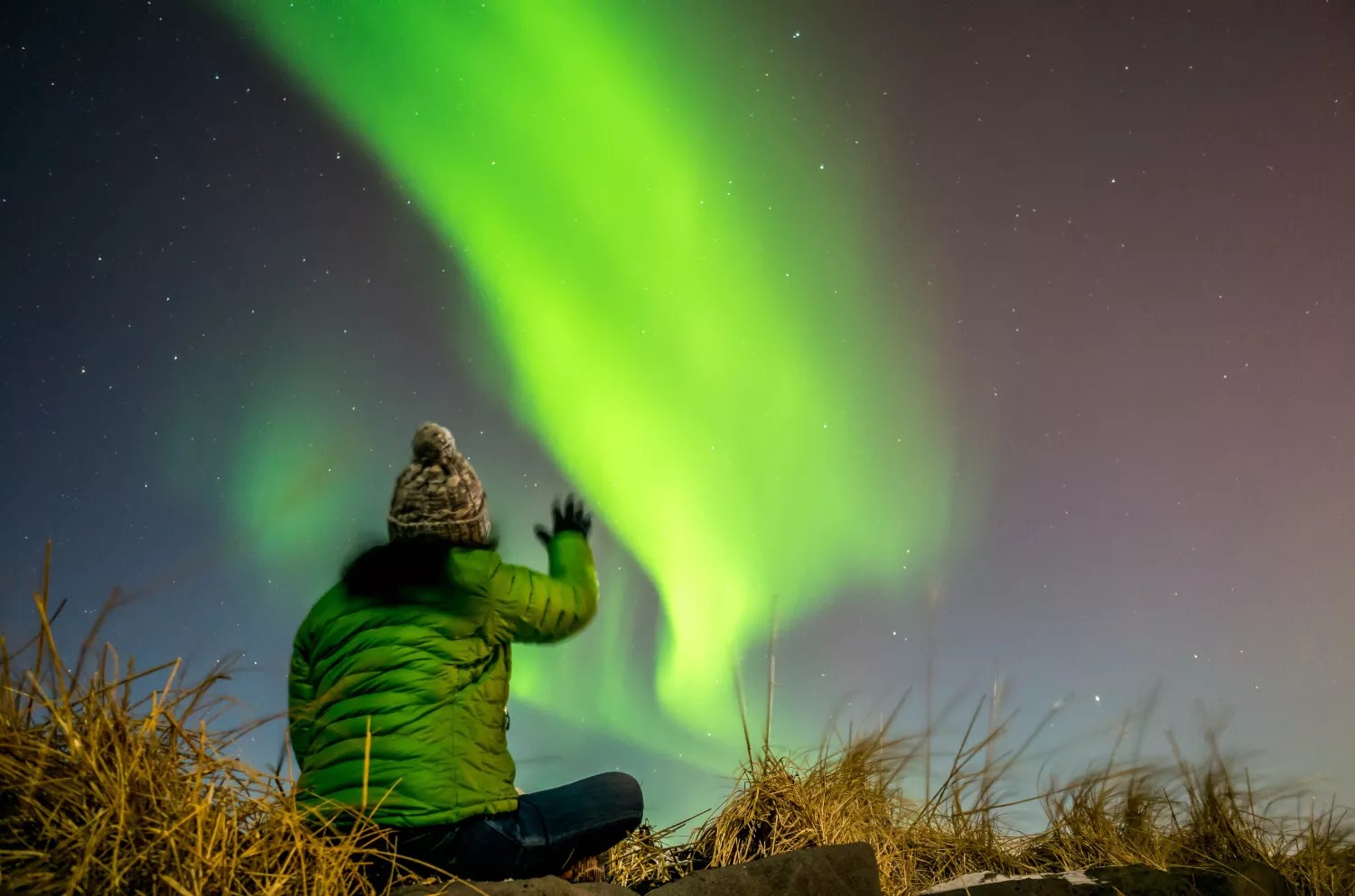 photo of a person in a beanie and thick green jacket looking up at green Northern Lights