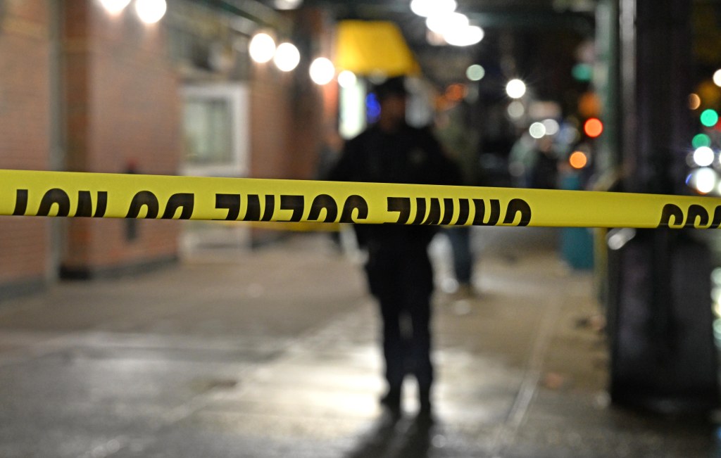 Yellow crime scene tape with black "DO NOT CROSS" text in front of a blurry street scene at night.