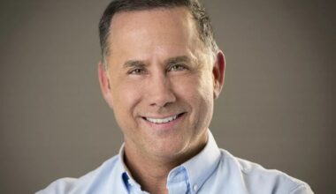 a whiteman in a light blue button-down smiles at the camera in front of a grey background