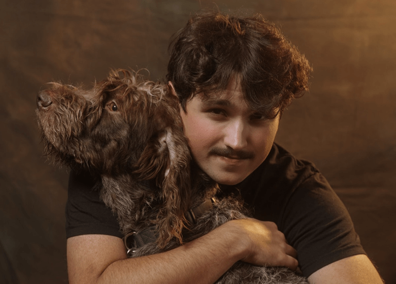 A high-contrast concert shot of a vocalist mid-performance on a dark stage. A single spotlight highlights the musician as they shout into the mic, with one hand visible on the fretboard of a guitar in the lower right corner.