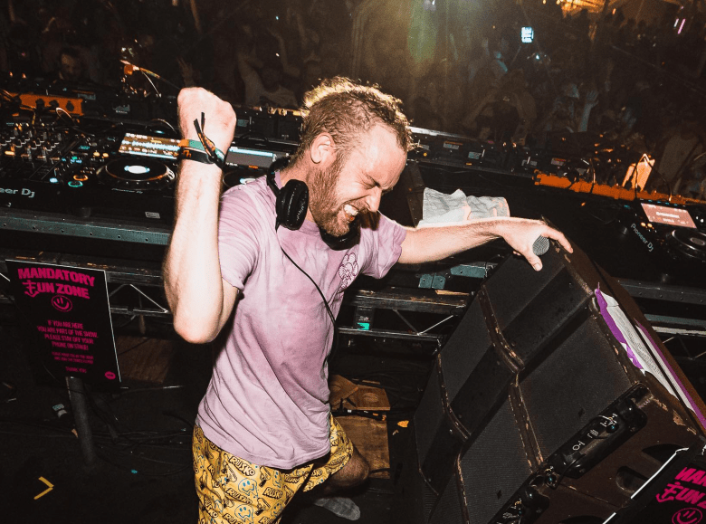 A musician with a short beard and headphones is performing at a music festival or concert. DJ equipment (Pioneer CDJs) is visible on the tables behind them, and warning signs reading "MANDATORY FUN ZONE" are visible on the speaker and surrounding area.