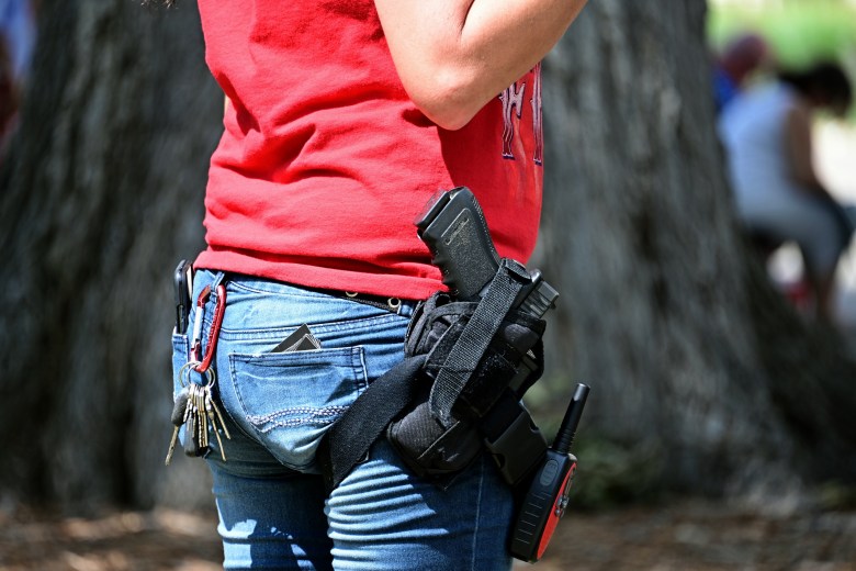 A close-up view from the waist down shows a person standing outdoors, wearing blue jeans and a red t-shirt. A black handgun is secured in a black thigh holster on their right leg, with a walkie-talkie clipped just below it. On the left side, a set of keys hangs from a red carabiner on a belt loop, and a card protrudes from the back pocket. The background is blurred, showing a large tree trunk and indistinct figures.