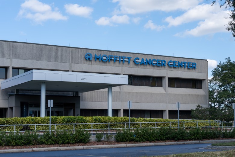 A daytime exterior view of the Moffitt Cancer Center. The large, beige concrete building features a covered entrance canopy with the number "4101" visible. Prominent blue lettering on the upper façade reads "MOFFITT CANCER CENTER." Green hedges and landscaping line the front of the building under a bright blue sky with scattered clouds.