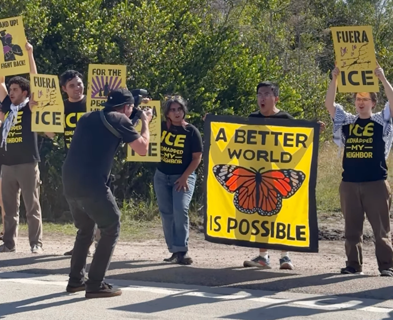 A group of protestors stands outside holding yellow and black signs. A cameraman films a man holding a large yellow banner featuring a monarch butterfly and the text, "A BETTER WORLD IS POSSIBLE."