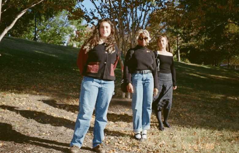 A full-length outdoor portrait of three individuals standing on a leaf-strewn grassy hill under the shade of trees. The person on the left wears a black and red jacket and blue jeans; the central person has blonde braids and wears a black turtleneck with jeans; and the person on the right wears an off-the-shoulder black top with a dark skirt.