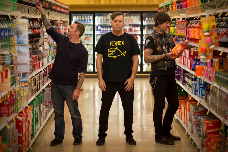 Three band members pose in the middle of a grocery store. The subject on the left is reaching for a beverage, and the person on the right is holding an orange juice container, both wearing denim. All three subjects have visible tattoos on their arms.
