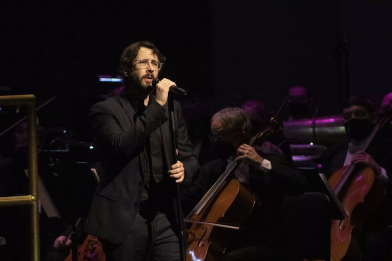 A high-contrast, dramatic photo of singer Josh Groban performing on stage. He is wearing a dark suit, a black shirt, and clear-rimmed glasses, holding a microphone close to his face with a focused expression. In the dimly lit background, several members of an orchestra are visible, including two cellists in the mid-ground. The lighting is moody, with purple and white highlights catching the performers against the dark stage.