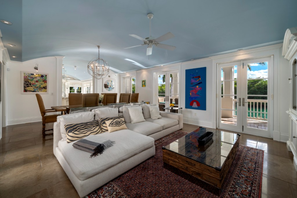 A living room with light blue ceilings, white walls, and french doors that lead to a patio overlooking the ocean