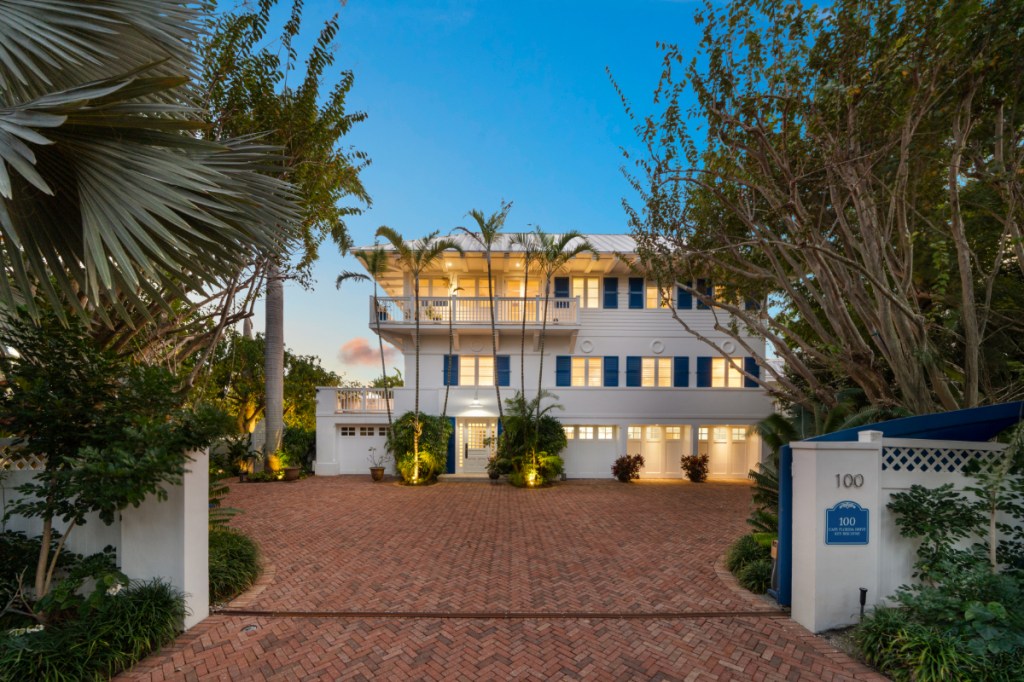 a large white home with blue shutters sits on a red brick-paved driveway, surrounded by greenery