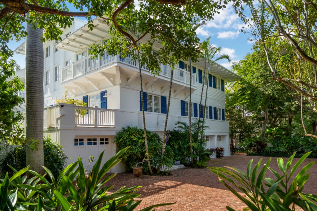 sideview of two-story white house in Key Biscayne. You see the driveway with trees surrounding the house. There are blue window shutters. 