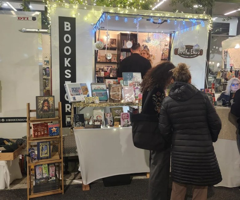 Customers browsing the 'Book Ends' mobile bookstore trailer, which is illuminated by festive string lights at a night market.