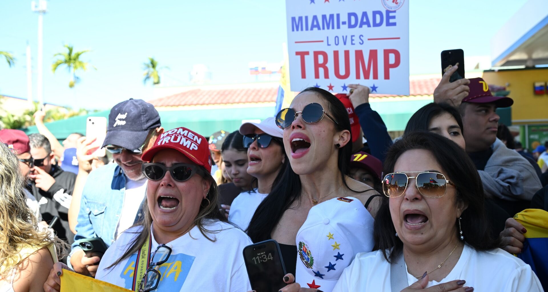 Photo of protesters singing at a rally. One woman wears a hat reading, "Women for Trump." A person in the back holds a sign reading, "Miami-Dade loves Trump"