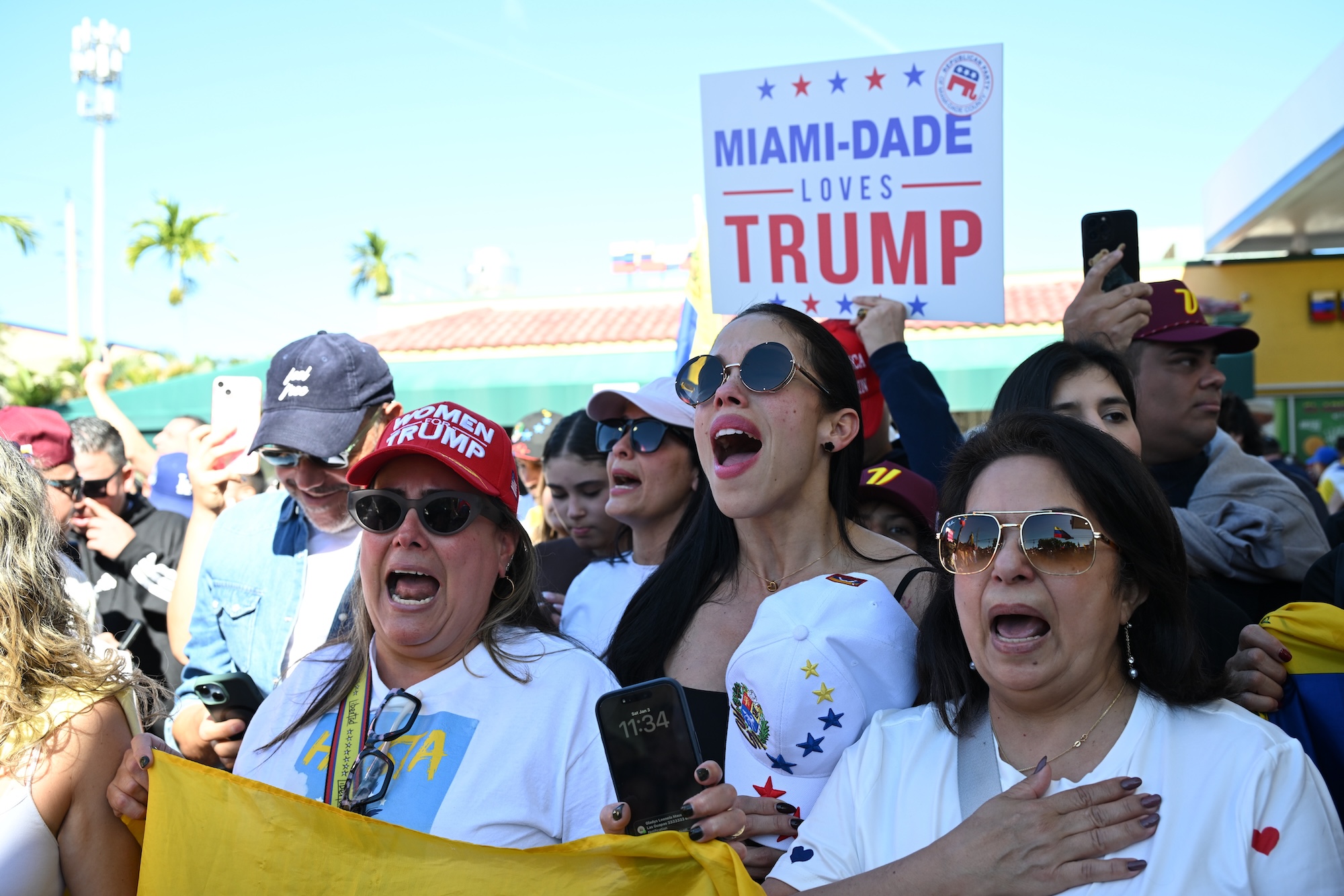Photo of protesters singing at a rally. One woman wears a hat reading, "Women for Trump." A person in the back holds a sign reading, "Miami-Dade loves Trump"