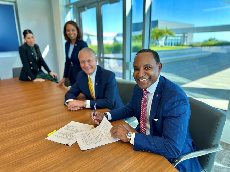 Michael Stephens signs papers sitting at desk with three other people behind him. 