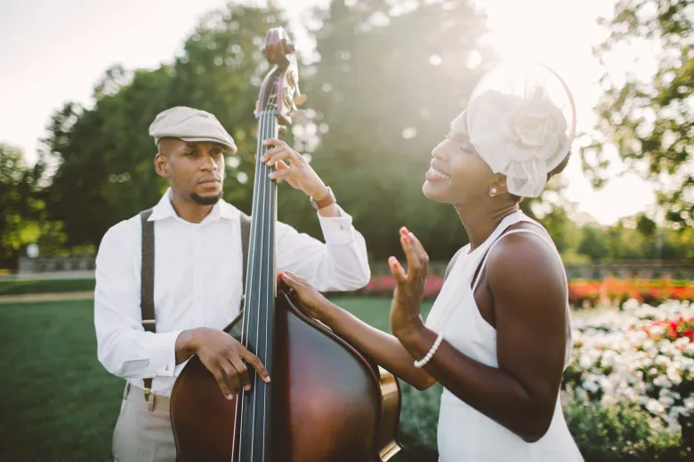 A sun-drenched outdoor portrait of the jazz duo Acute Inflections. On the left, bassist Sadiki Pierre, a Black man wearing a tan flat cap, white button-down shirt, and dark suspenders, looks focused as he plays a polished wooden upright bass. On the right, vocalist Elasea Douglas, a Black woman in a white sleeveless dress and a white floral fascinator, smiles with her eyes closed, gesturing gracefully with one hand as if singing. They are positioned in a lush green park with a soft-focus background of trees and colorful flower beds, captured in warm, glowing natural light.