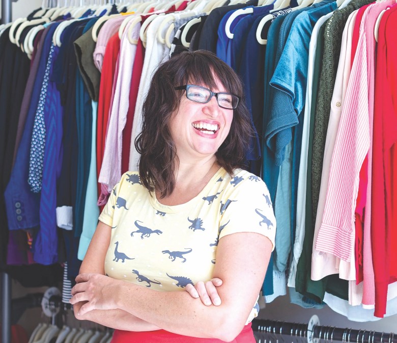 A smiling woman wearing a dinosaur print t-shirt standing in front of racks of colorful clothing.