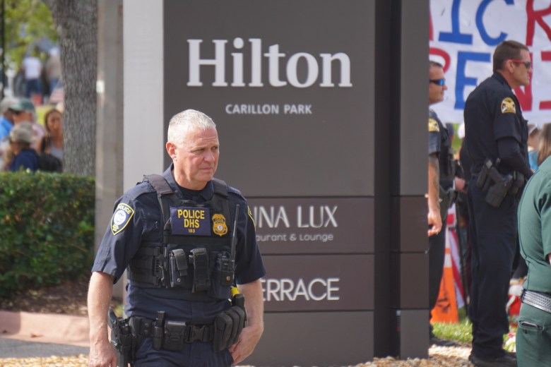 Law enforcement officers wearing tactical vests stand near a Hilton Carillon Park sign during an outdoor event, with people and protest signage visible in the background.