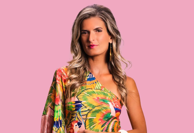 A studio portrait of a person with long, wavy blonde hair and pink lipstick, posed against a solid pink background. They are wearing a vibrant, one-shoulder tropical print garment featuring green palm leaves and orange-and-yellow Macaw patterns, finished with fringe on the sleeve.