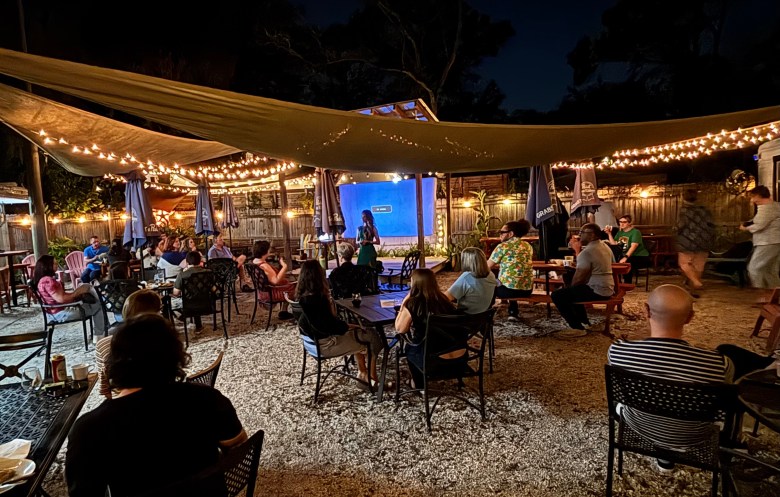 A crowd of people seated at an outdoor patio at night, listening to a speaker on stage under warm string lights.
