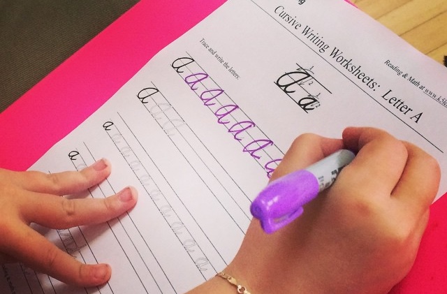 A child's hand holds a purple marker writing the letter "a" multiple times on a cursive practice sheet.