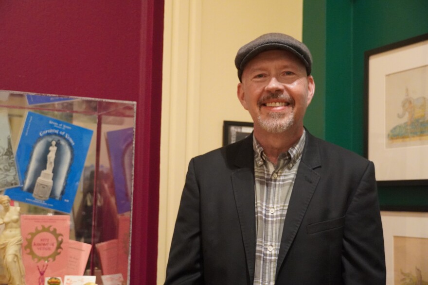 Man in black jacket and plaid shirt and fedora smiles at camera with artifacts behind him 