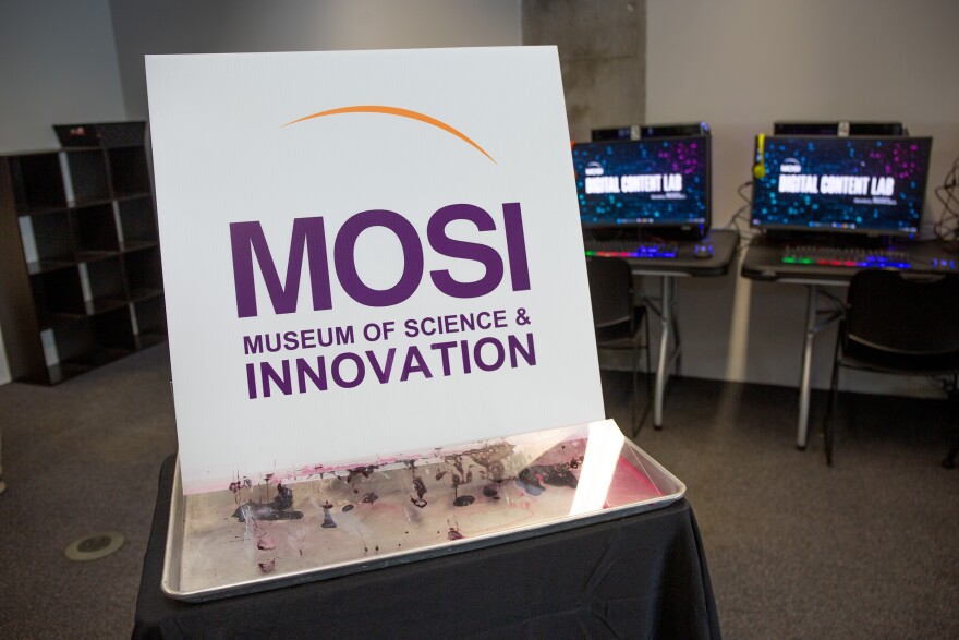 A close up of MOSI's new name on a white sign. The Museum of science and industry. It rests on a metal plate holding the remnants of a foam card that said "industry," which was melted using acetone to reveal the new name - innovation.
