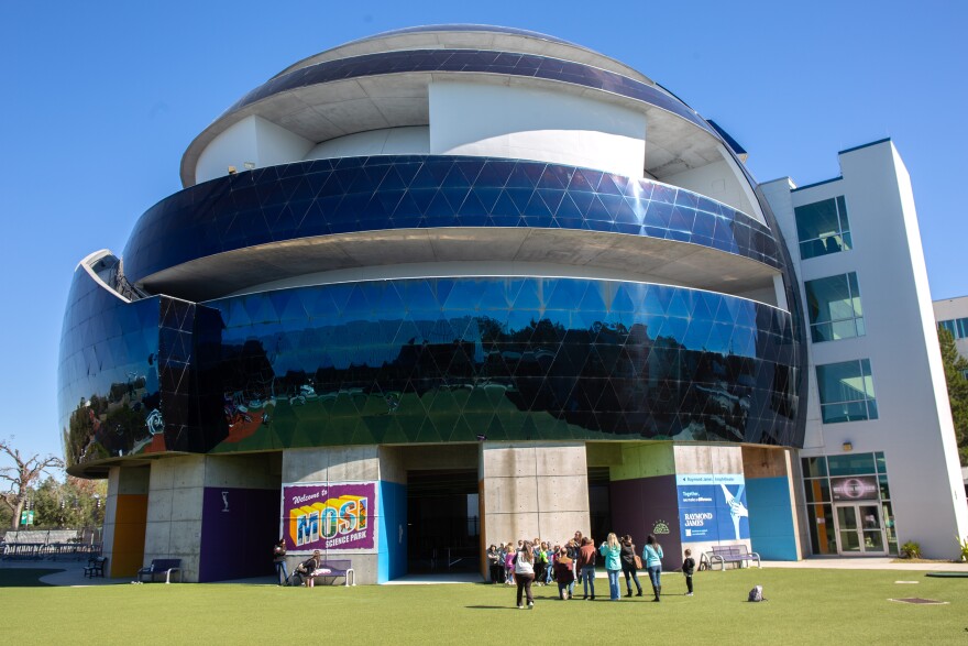 The outside of a large, circular dome made of concrete and blue glass panels. Students are milling about on the lawn in front of it. 