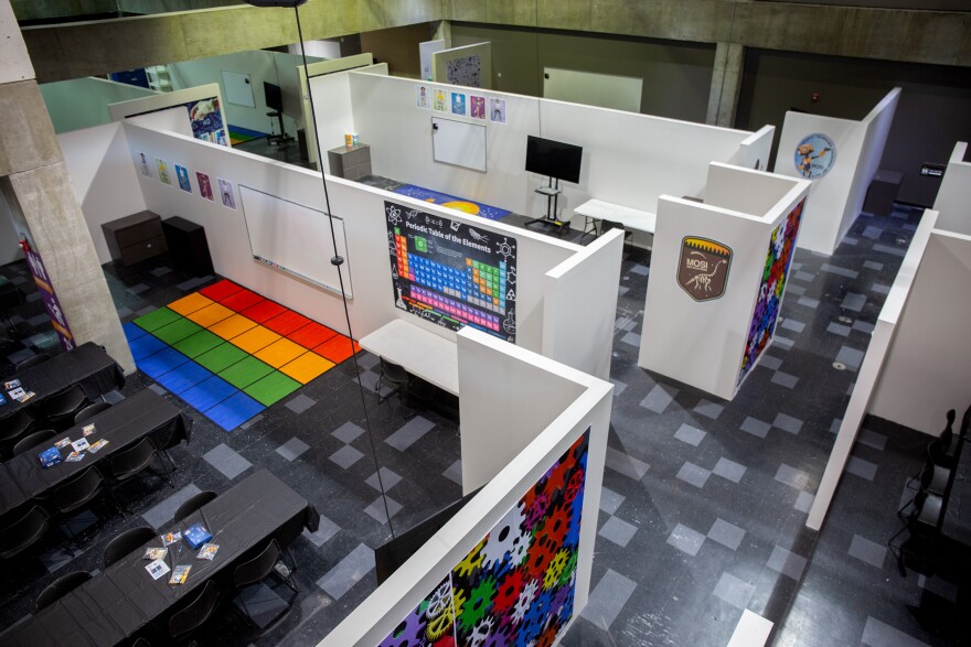An overhead view of a long stretch of flooring, with cubicle-like walls sectioning off areas into classrooms. In one classroom, there is a row of tables, a colorful rug, and the periodic table of the elements. 