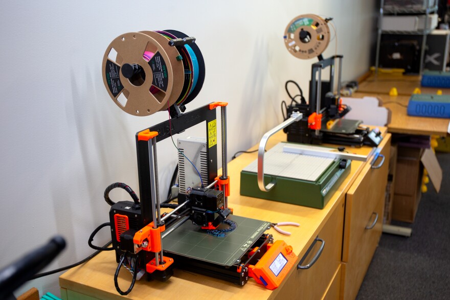 A close up of two black and orange, small-scale 3D printers. Both are feeding resin from coils mounted above them to produce items. 