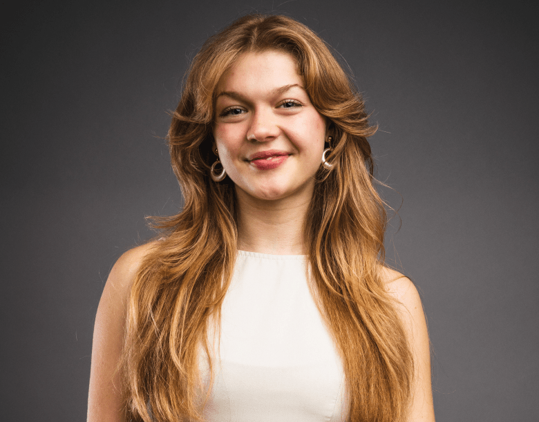 A studio portrait of a person with long, flowing strawberry-blonde hair and a friendly smile. They are wearing a sleeveless white top and large gold hoop earrings, posed against a solid dark grey background.