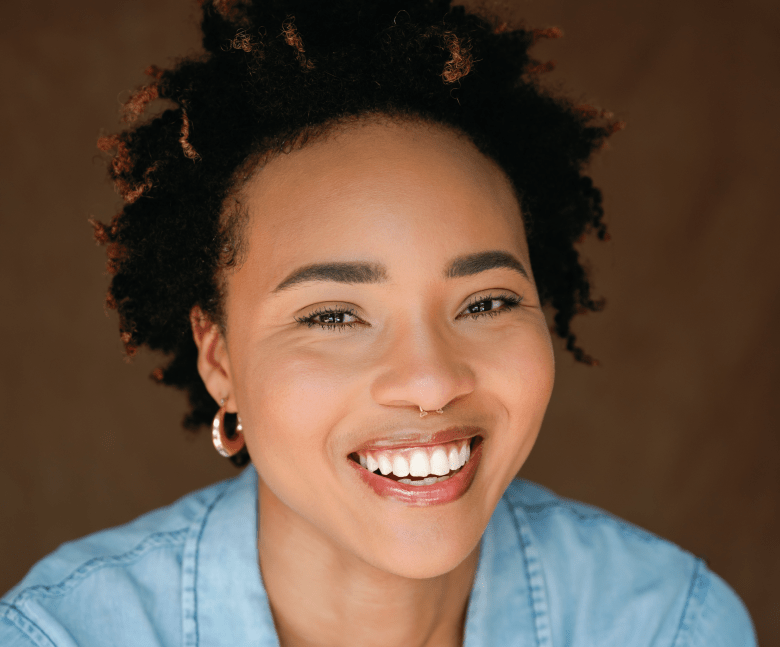 A tight, vibrant headshot of a person with a joyful expression and a wide smile, framed against a warm brown background. They have short, voluminous dark hair with light brown highlights and are wearing a light blue button-down shirt, a small gold hoop earring, and a subtle gold nose ring.