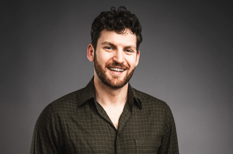 A studio portrait of a person with short, curly dark hair and a beard, looking toward the camera with a wide smile. They are wearing a dark olive-green button-down shirt with a subtle grid pattern, posed against a solid grey background.