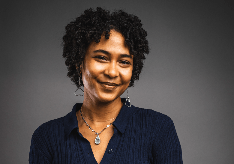 A studio portrait of a person with short, curly dark hair and a warm, closed-mouth smile. They are wearing a navy blue ribbed polo shirt, heart-shaped silver drop earrings, and several layered necklaces—including one with a teardrop-shaped pendant—all set against a solid grey background.