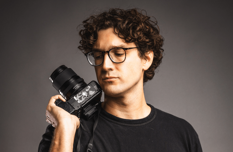A studio portrait of a person with curly brown hair and tortoise-shell glasses, looking down thoughtfully at a professional camera they are holding against their shoulder. They are wearing a black crew-neck t-shirt, posed against a solid dark grey background.