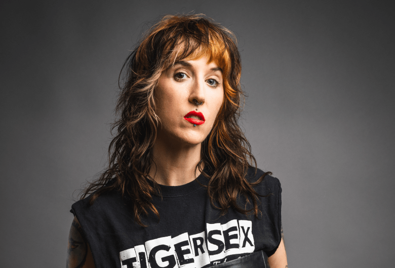 A studio portrait of a person with long, wavy brown hair featuring blonde highlights and a shaggy fringe. They are wearing a black "TIGERSEX" sleeveless t-shirt, bright red lipstick, and multiple facial piercings, including a septum ring and a lip piercing, all set against a solid grey background.