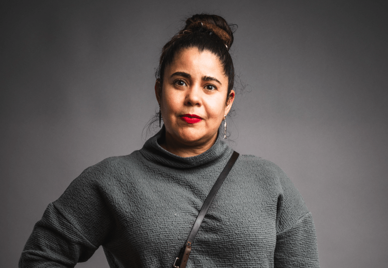 A studio portrait of a person with dark hair styled in a high bun, looking directly at the camera with a neutral expression. They are wearing a grey textured turtleneck sweater, bright red lipstick, and small silver fish-shaped earrings, all set against a solid grey background.