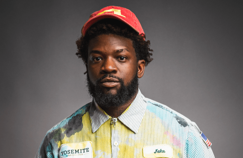 A studio portrait of a person with a dark beard and a red baseball cap, looking directly at the camera with a neutral expression. They are wearing a unique, multi-colored button-down shirt featuring yellow and blue tie-dye patterns, a "Yosemite National Park" patch, a "Jake" name tag, and an American flag patch on the sleeve, all set against a solid grey background.