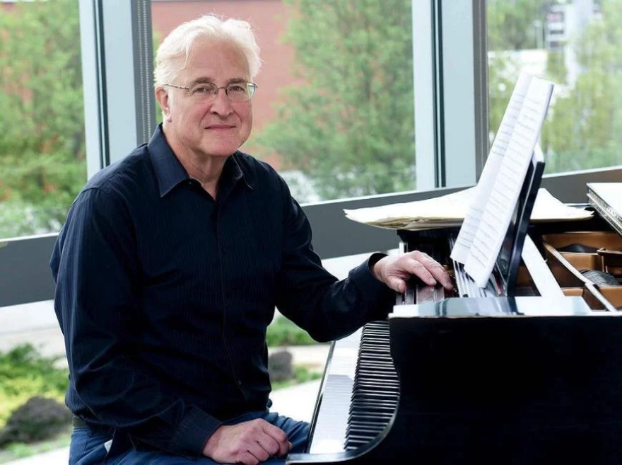 A mane in a dark shirt sits at a grand piano. He has snowy white hair and glasses.