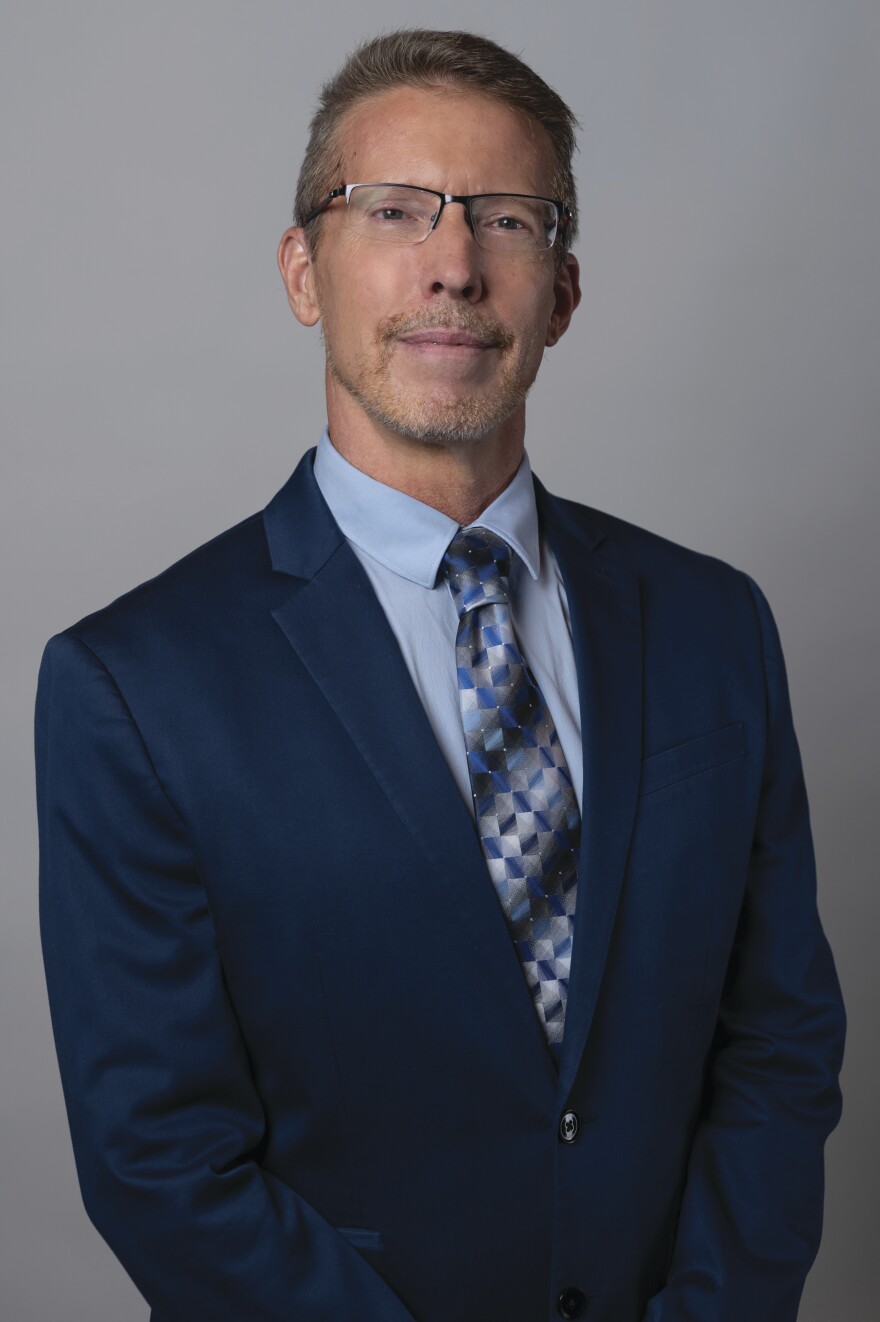 A man with close-cropped hair and glasses with a scruffy beard, wearing a navy suit with a blue shirt and geometric tie, looks at the camera. 